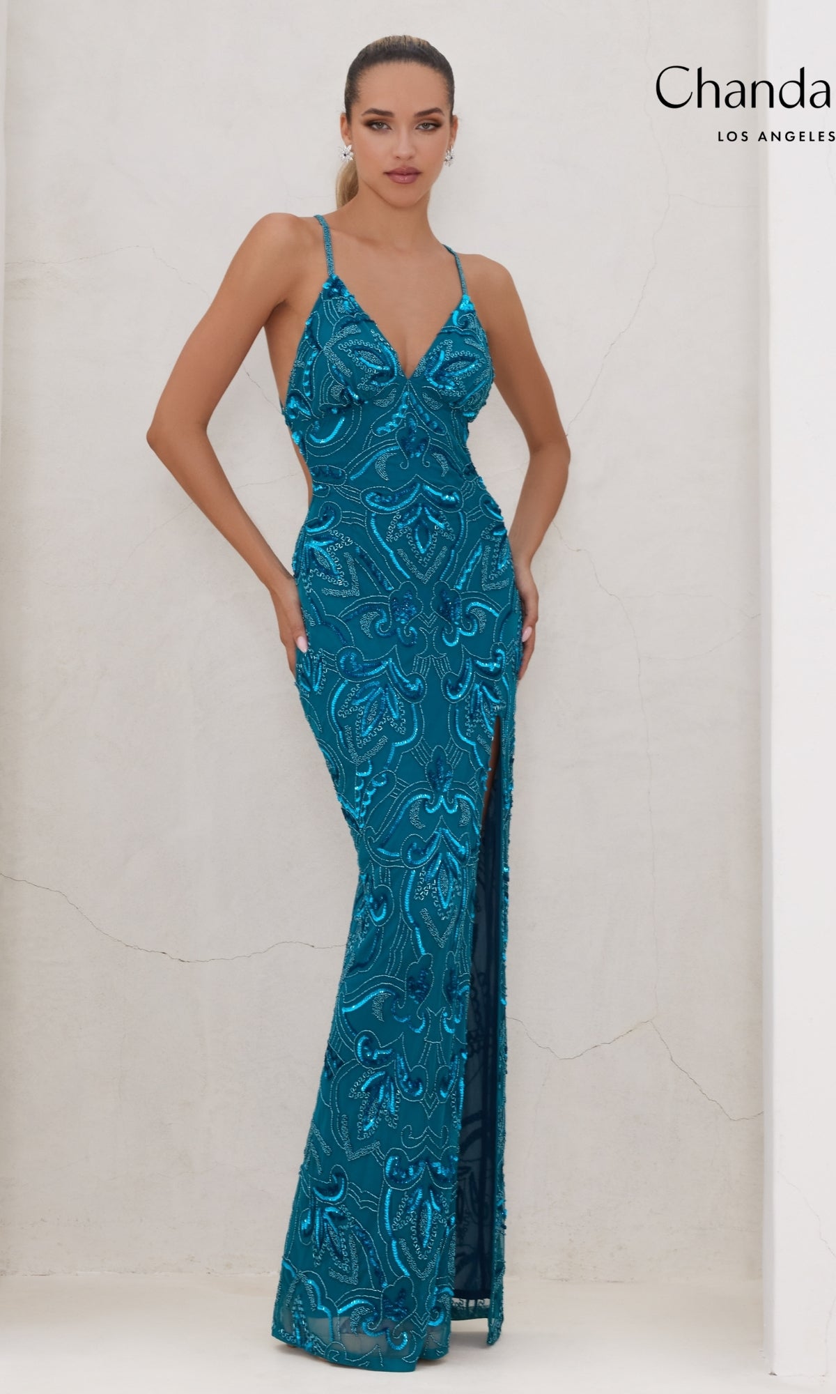 A woman models the Long Prom Dress 30020 by Chandalier—a teal, sleeveless, floor-length sequin formal dress with a deep V-neckline and thigh-high slit—standing against a light-colored wall.